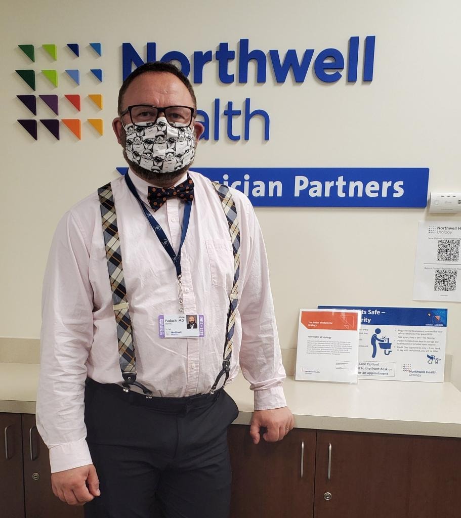 A doctor in suspenders stands in front of a Northwell health sign.